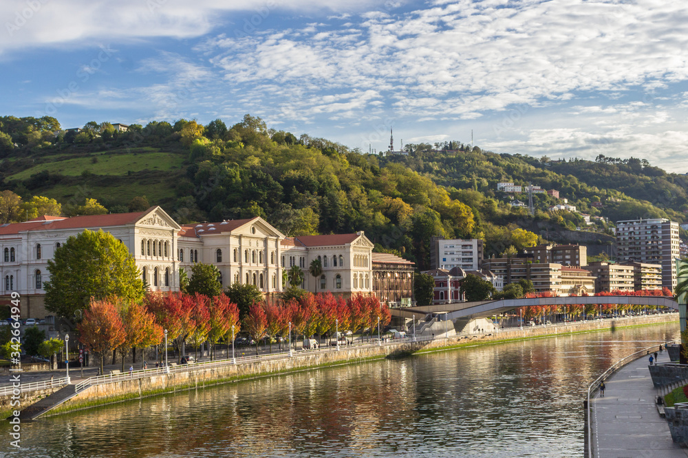 Fototapeta premium Embankment by the Nervion River in Bilbao. Bend of the river. Colors of autumn in the city. Traveling in Spain. European city. Colorful cityscape by the river. Basque Country. The large city of Spain.