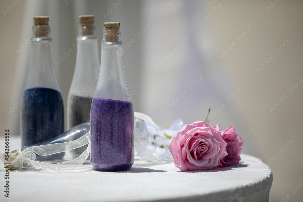 Wedding decor of bottles filled with colored sand  and ribbon and pink rose for decoration on a table with white linen