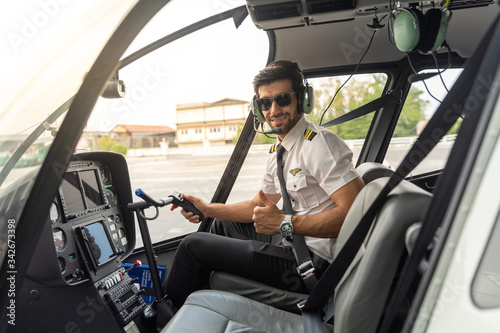 Picture of a male helicopter pilot in his white shirt with tie uniform. He is prepared for his flight. He sits inside a white helicopter with his head phone one ready to take off.