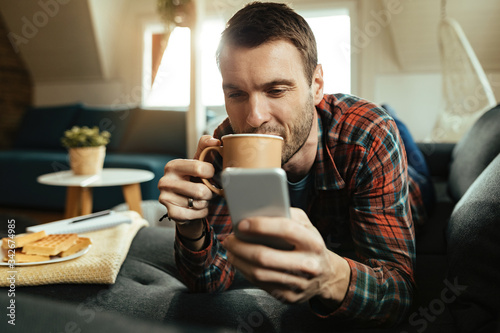 Young man drinking coffee and using smart phone while relaxing at home.