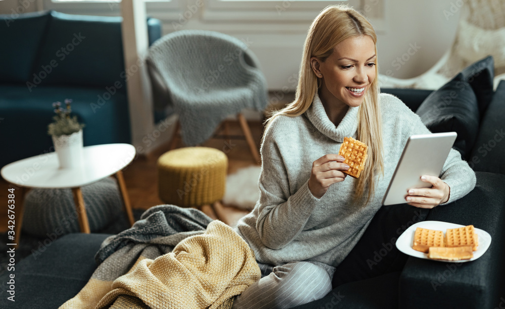 © Drazen - Happy woman using touchpad and eating waffles in the living room.