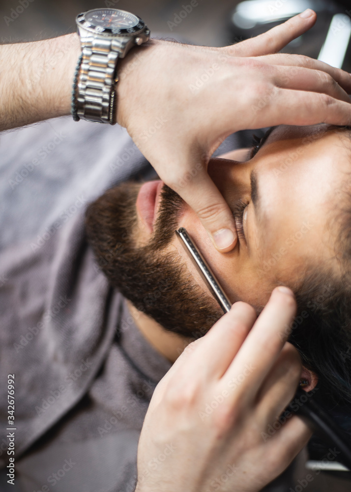 
Barber hands with a razor shaving beard of male customer