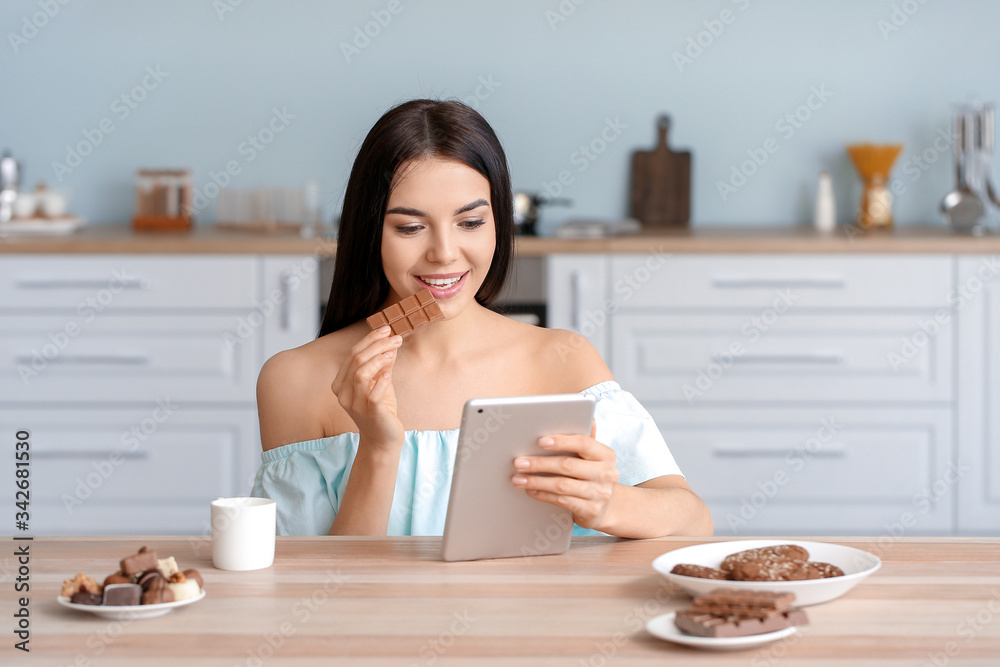 Beautiful young woman with tablet computer eating chocolate in kitchen Beautiful young woman with tablet computer eating chocolate in kitchen