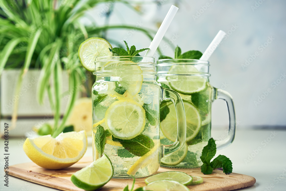 Two glasses of homemade lemon, lime, and mint lemonade sit on the wooden dining table. Cold, refreshing summer lemonade or mojito. - obrazy, fototapety, plakaty 