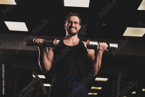 A man with an effort does an exercise for biceps and triceps with a dumbbell. Embossed bodybuilder smiles. Muscular guy with glasses in the gym.