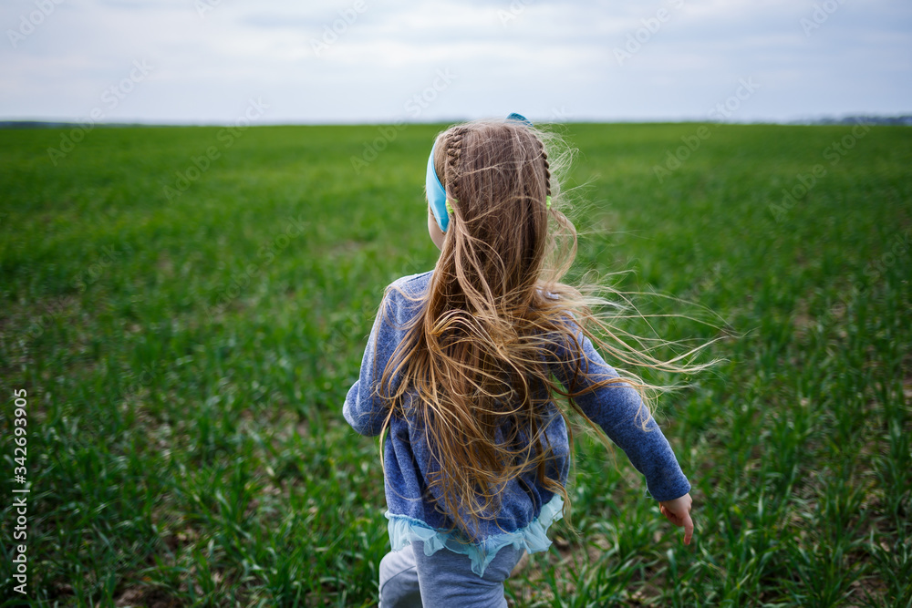 Fototapeta premium little girl child runs on green grass in the field, sunny spring weather, smile and joy of the child, blue sky with clouds