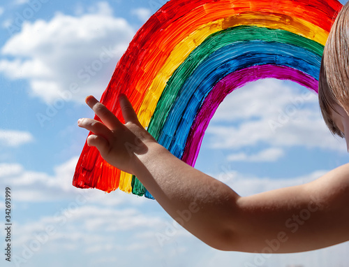 the child points to the painted rainbow symbol on the window. blue sky with clouds. symbol of happiness and love