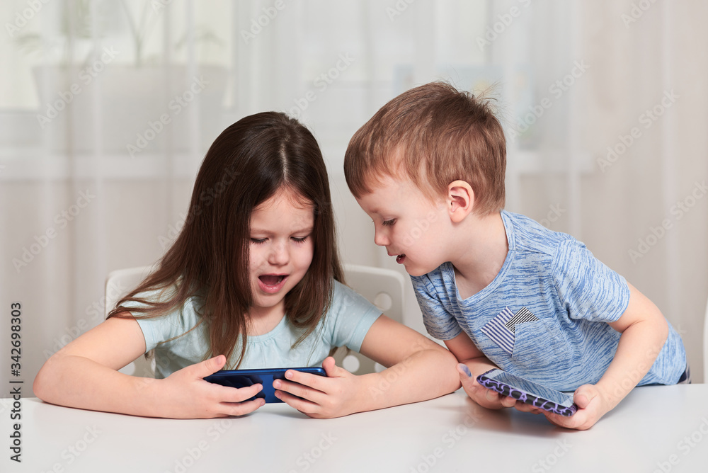 A little girl and a boy sit at a table and watch a video on a tablet. A little girl and a boy sit at a table and watch a video on a tablet.