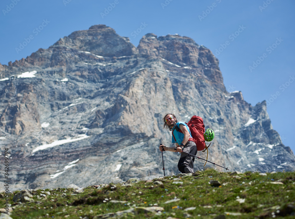 Smiling traveler with backpack walking rocky hillside path. Hiker with trekking sticks traveling alone in mountainous region, climbing on Matterhorn or Monte Cervino. Concept of hiking, mountaineering