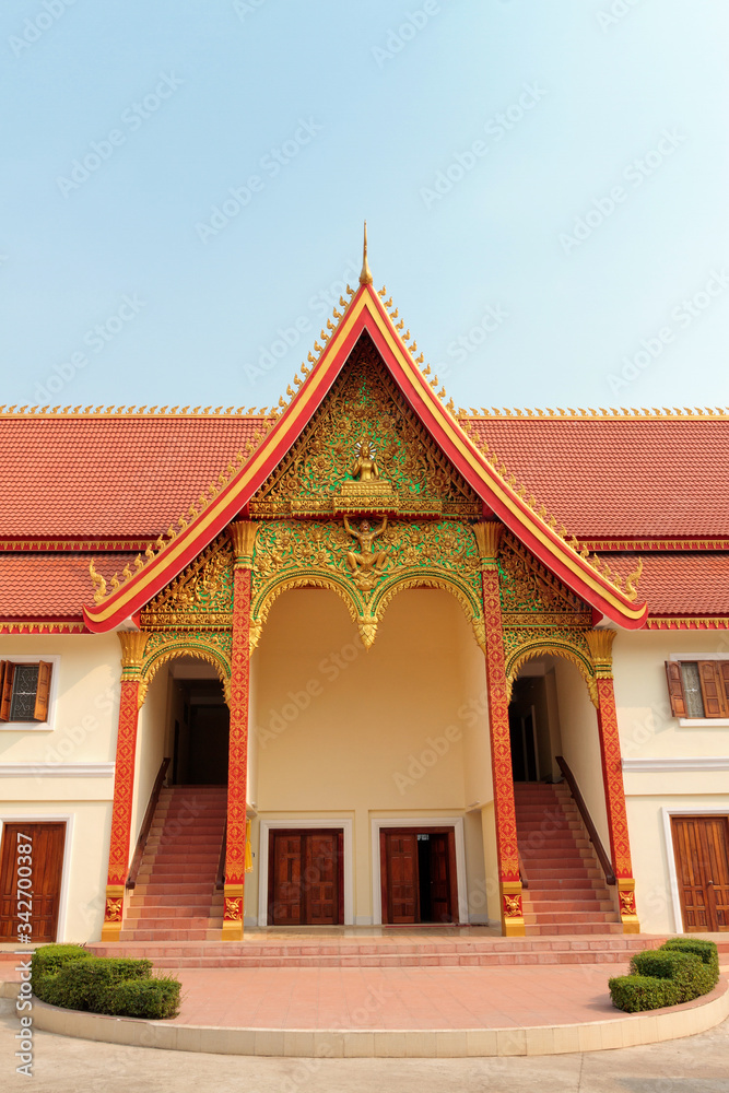 An entrance of Buddhist temple with ornate red roof with golden decoration in Vientiane in Laos against blue sky