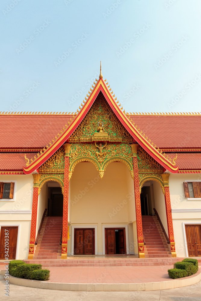 An entrance of Buddhist temple with ornate red roof with golden decoration in Vientiane in Laos against blue sky An entrance of Buddhist temple with ornate red roof with golden decoration in Vientiane in Laos against blue sky