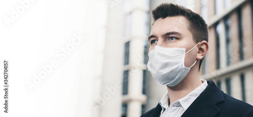 Businessman and coronavirus epidemic. Close up portrait of young business man in a disposable facial mask. A man defends himself against covid 19 on the big city street. Modern buildings at background