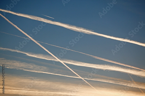 A plane is flying across a morning sky that has many Chemtrails from travelling on a blue sky at sunrise.