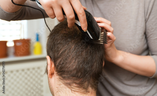 Woman is doing haircut with hair trimmer to man at home.