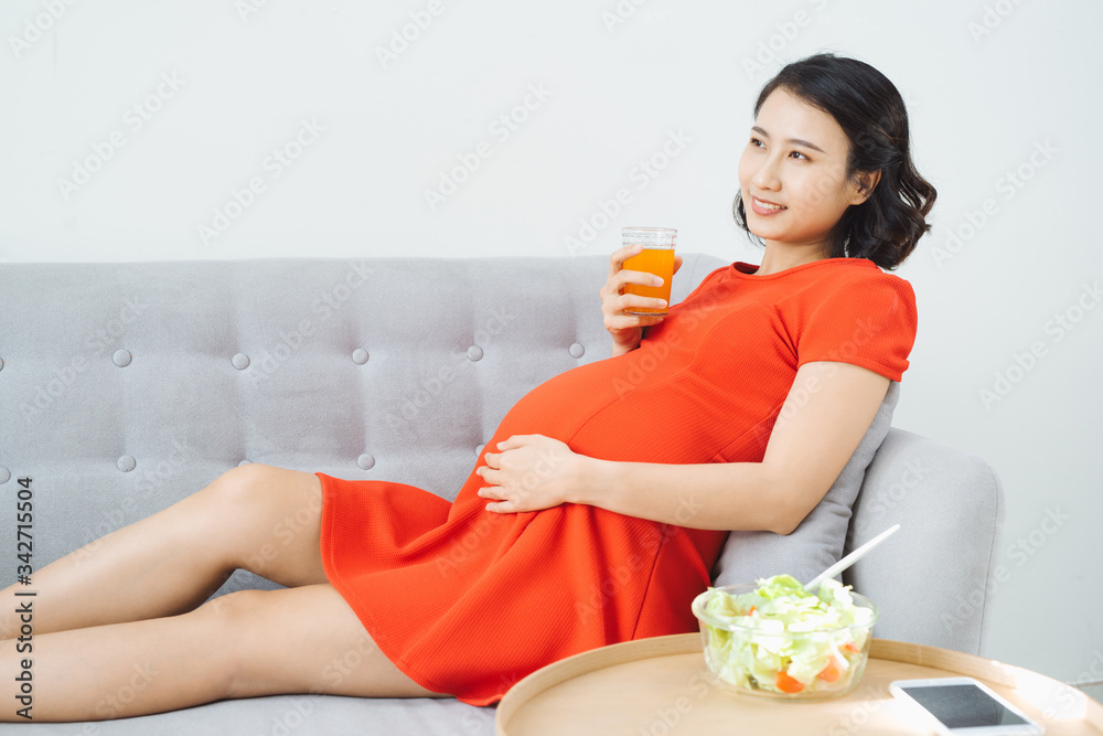 Pretty young Asian pregnant woman eating salad and drinking milk when sitting on sofa.