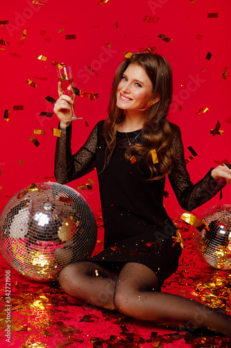 Brunette girl sits in a black dress, holds a glass of champagne in her hands, fly gold confetti on a red background