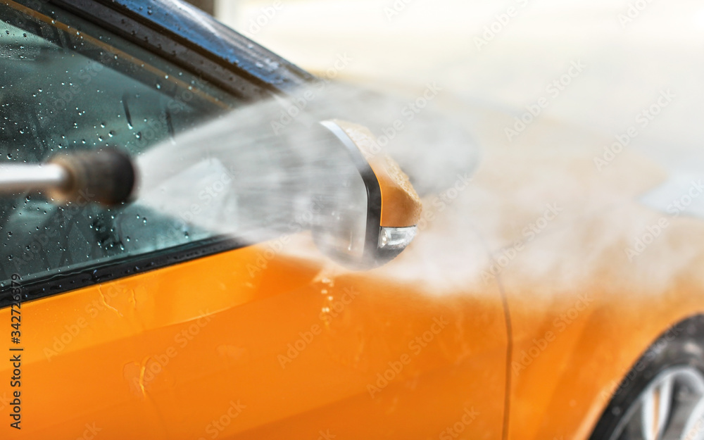 Side mirror of yellow car being washed in self service carwash, water spraying with high pressure, drops flying around.
