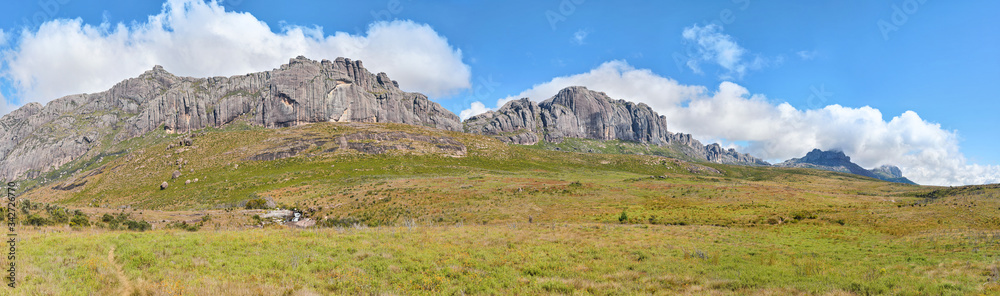 Wide panorama of grass fields, Andringitra massif tall rocky mountains in distance, blue sky above. Typical scenery seen during trek to Pic Boby peak