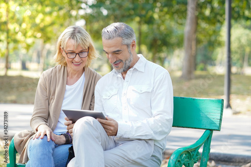 Elderly mature senior couple sitting at bench in the park and looking at tablet or smart devices to connect internet social media watching movie and video chat with happy and enjoy lifestyle