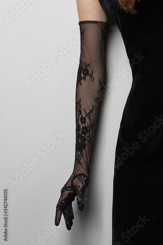 Cropped shot of a lady in a black tight dress and a black evening  glove against the gray wall. The elegant elbow glove is made of the lace fabric with floral pattern. 