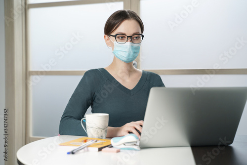 A young woman in a medical mask works on a laptop at home office