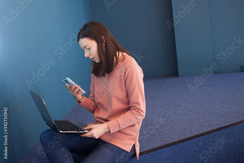 student sits on the steps in the recreation area and uses a laptop and a smartphone, a young woman