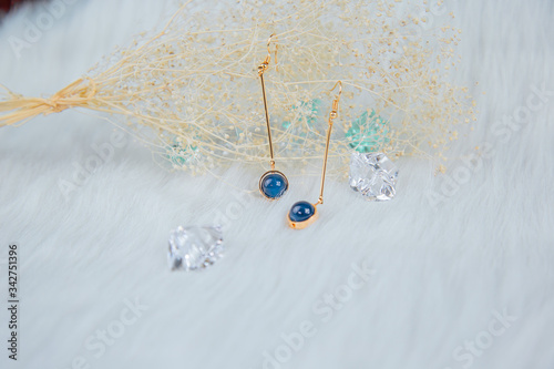Earrings made of blue round beads and golden yellow metal. Yellow dry grass plants as a background. White plush cloth and transparent ice ornaments.