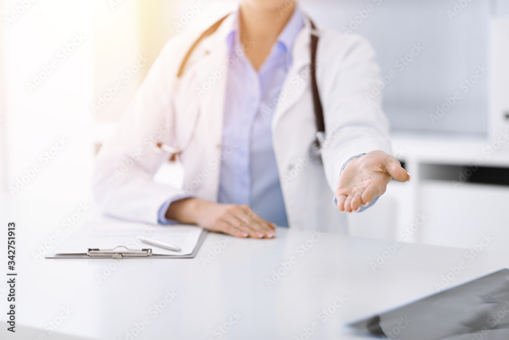 Unknown woman-doctor in blue blouse is giving helping hand while sitting at the desk in sunny clinic. Medicine concept Unknown woman-doctor in blue blouse is giving helping hand while sitting at the desk in sunny clinic. Medicine concept
