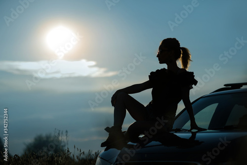 Silhouette of a girl in sunglasses sitting on the hood of a car against the sunset sky.