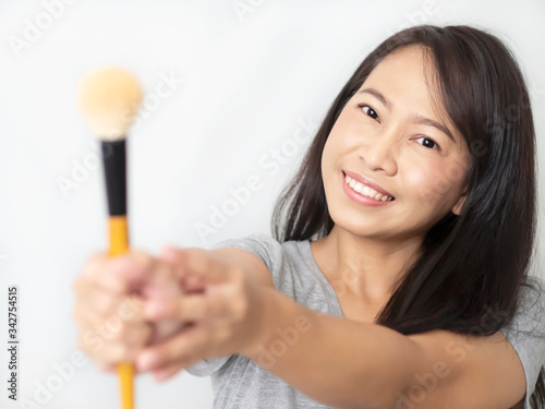 Asian women wearing gray T-shirts are smiling happily. Lady is holding make-up brush extending to camera. Female has dark brown long hair is cheerful and make up pink nude tone color. White background
