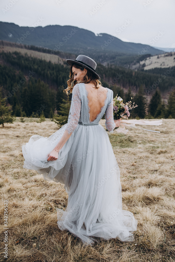 Portrait of the bride. A young girl in a blue gray wedding dress and hat with a bouquet of flowers and greenery in her hands on a background of mountains and a forest at sunset