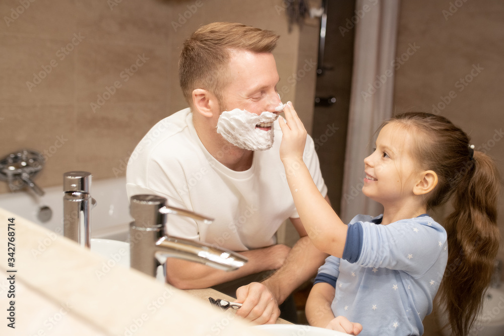 Happy cute little girl touching her nose of her father with shaving foam on face