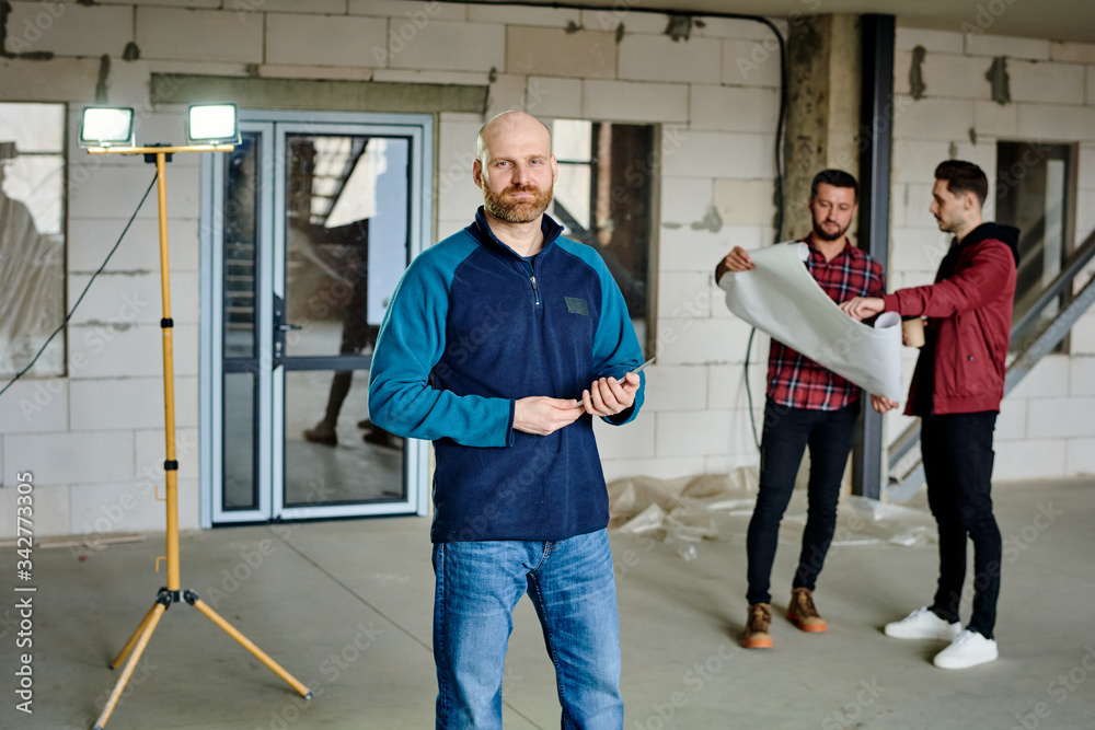 Confident foreman with tablet standing in front of camera in unfinished building Confident foreman with tablet standing in front of camera in unfinished building