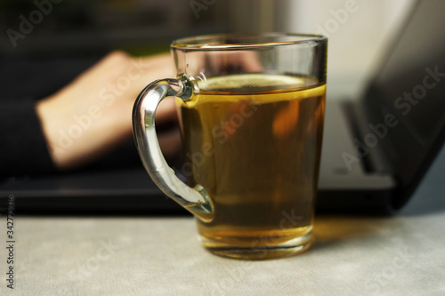 Young woman working on a laptop on the kitchen table, remote work at home, self-isolation, selective focus on a Cup of tea with lemon