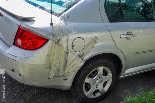 Damaged small gray car parked on a residential street. The rear panel is held on to car with adhesive tape