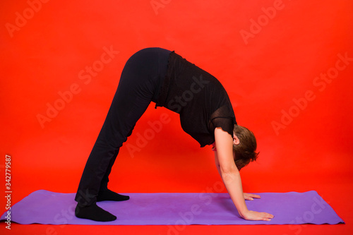 Aged woman in black clothes doing yoga on a purple rug. Studio photo on a red background. 