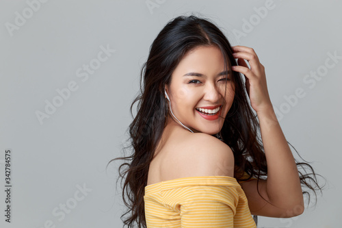 Close up young attractive Asian woman wearing yellow t shirt, earphones listening music with smiley face, studio head shot isolated on grey. photo