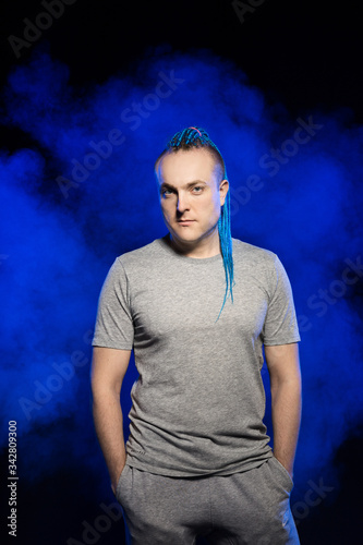 A man with a braided mohawk haircut calmly posing holding hands in pockets of sweatpants against a background of blue-red smoke.