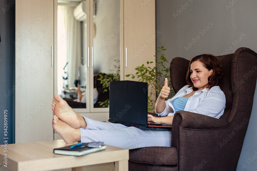 A 30-year-old young woman in a white shirt sits at home in a comfortable armchair and communicates with relatives using a laptop and video calling. A 30-year-old young woman in a white shirt sits at home in a comfortable armchair and communicates with relatives using a laptop and video calling.