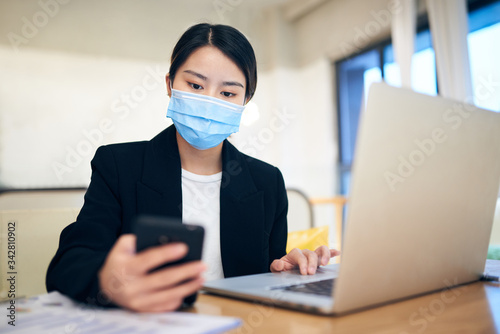 Young Asian woman wearing a face mask was using phone and laptop at home.