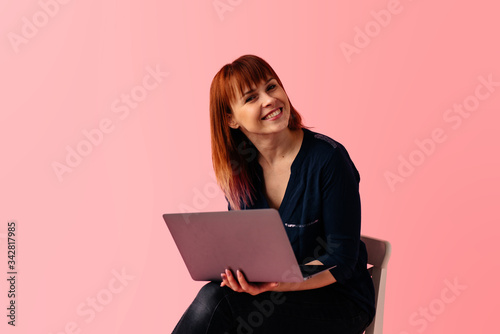 Young businesswoman working on the laptop sitting on tha chair in studio isolated on pink background