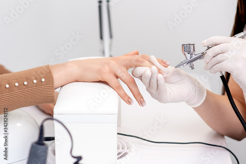 Manicure master is painting finger nails in beige color using airbrush.