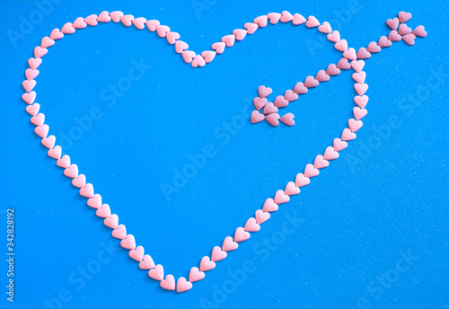 Heart shape pierced by arrow made from litle pink candy hearts on blue background