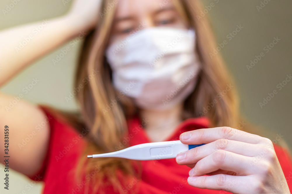 Close-up portrait of a woman holding her head with her hand and holding an digital thermometer in the other hand. Concept of illness, fever. Selective focus, blurred background.
