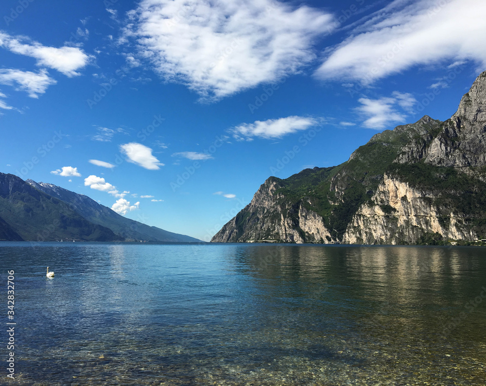 View from the lakeside of Riva del Garda in Italy looking out over the crystal clear waters of the lake, with tall mountains in the background