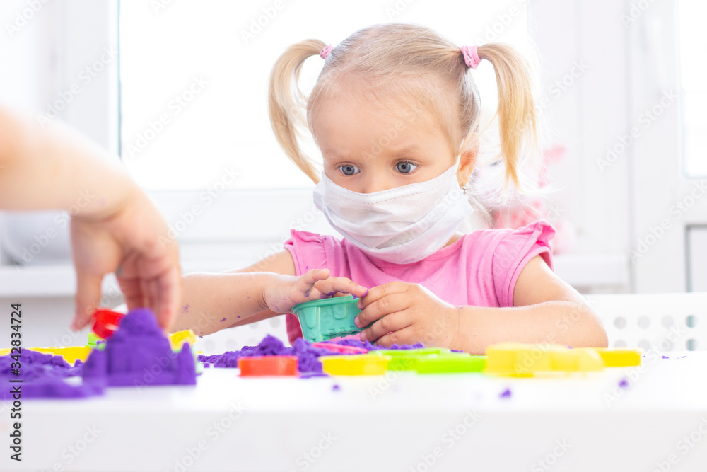 girl in a protective medical mask plays kinetic sand in quarantine. Blond beautiful girl smiles and plays with purple sand on a white table. coronavirus pandemic girl in a protective medical mask plays kinetic sand in quarantine. Blond beautiful girl smiles and plays with purple sand on a white table. coronavirus pandemic
