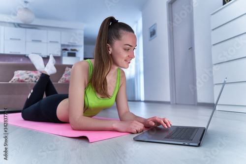Young happy attractive athlete woman in sportswear watches online training exercises and fitness workout on a computer on a mat at home gym