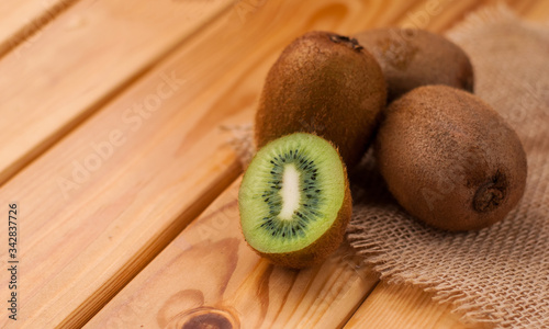 a green cut kiwi lies on a piece of burlap on a wooden table. there are several whole fruits in the back. banner, copyspace.