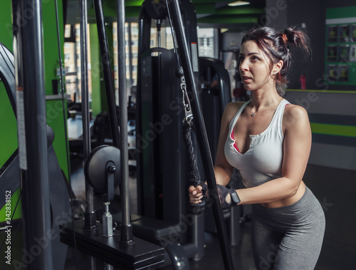 Young slim woman practicing extension of arms with ropes in exercise machine at gym