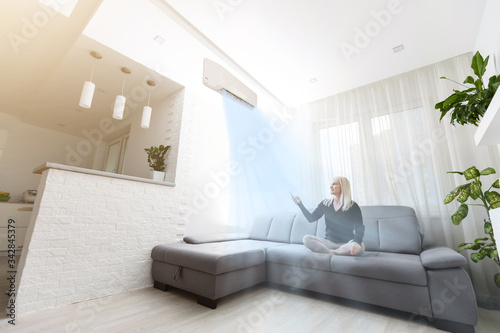 Young woman switching on air conditioner while sitting on sofa near white wall